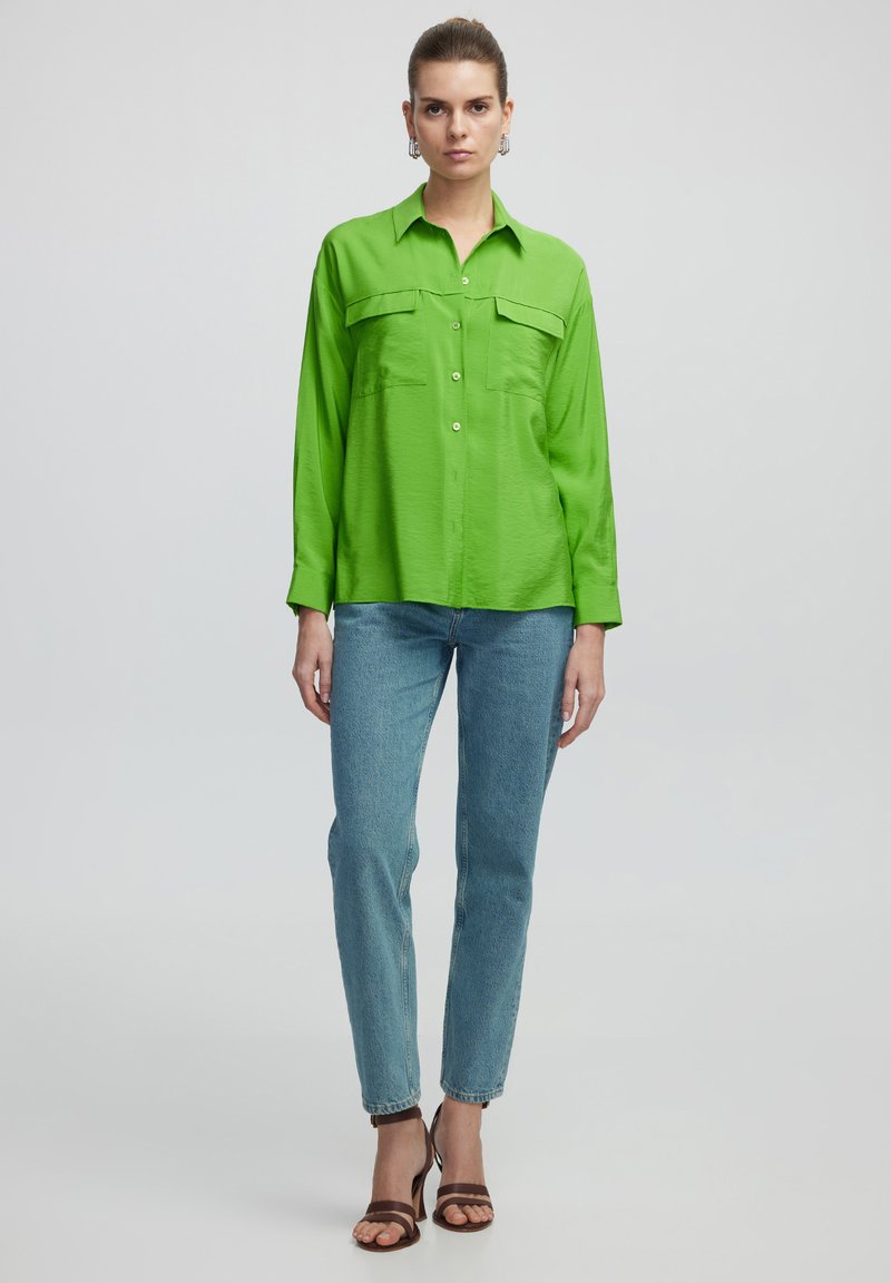 Zielona koszula zapinana na guziki z dwiema kieszeniami na piersi, długimi rękawami i luźnym krojem, zestawiona z jasnoniebieskimi dżinsami i brązowymi sandałami na obcasie.