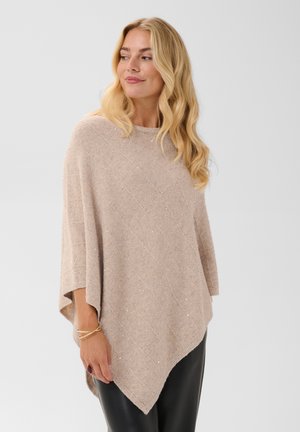 Femme aux longs cheveux blonds portant un poncho beige scintillant et un pantalon noir, regardant vers sa gauche avec un léger sourire.