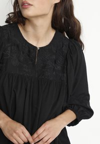 Svart blus med blommönsterbroderi vid halsringningen, plisserad bodice och puffärmar. Har en liten knapp vid framsidan.