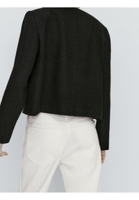 Svart kort jacka med textilier, öppen framtill och lång ärm. Kombinerad med ljusa jeans med bakfickor.