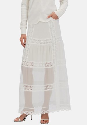 Weißer knöchellanger Rock mit Spitzen-Einsätzen, transparentem Stoff, getragen mit weißen High-Heel-Sandalen und einem langärmeligen Oberteil.