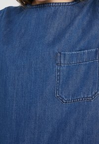 Tummansininen farkkupaita, jossa pyöreä pääntie, lyhyet hihat ja vasemmassa rinnassa tasku, jossa on hillitty tikattu reunus.