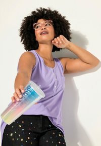 Top de tirantes morado con botones, combinado con unos jeans negros decorados con acentos coloridos en forma de joyas. Sostiene un vaso texturizado con un degradado.