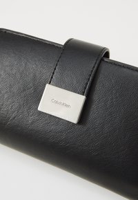 Cartera de piel negra con textura suave, que cuenta con una placa metálica plateada con el logo de Calvin Klein y un cierre de solapa.