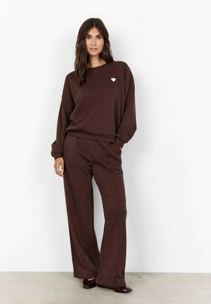 Kvinde iført en matchende mørkebrun sweatshirt og vidde bukser med et lille hvidt hjerte på brystet, stående mod en hvid væg.