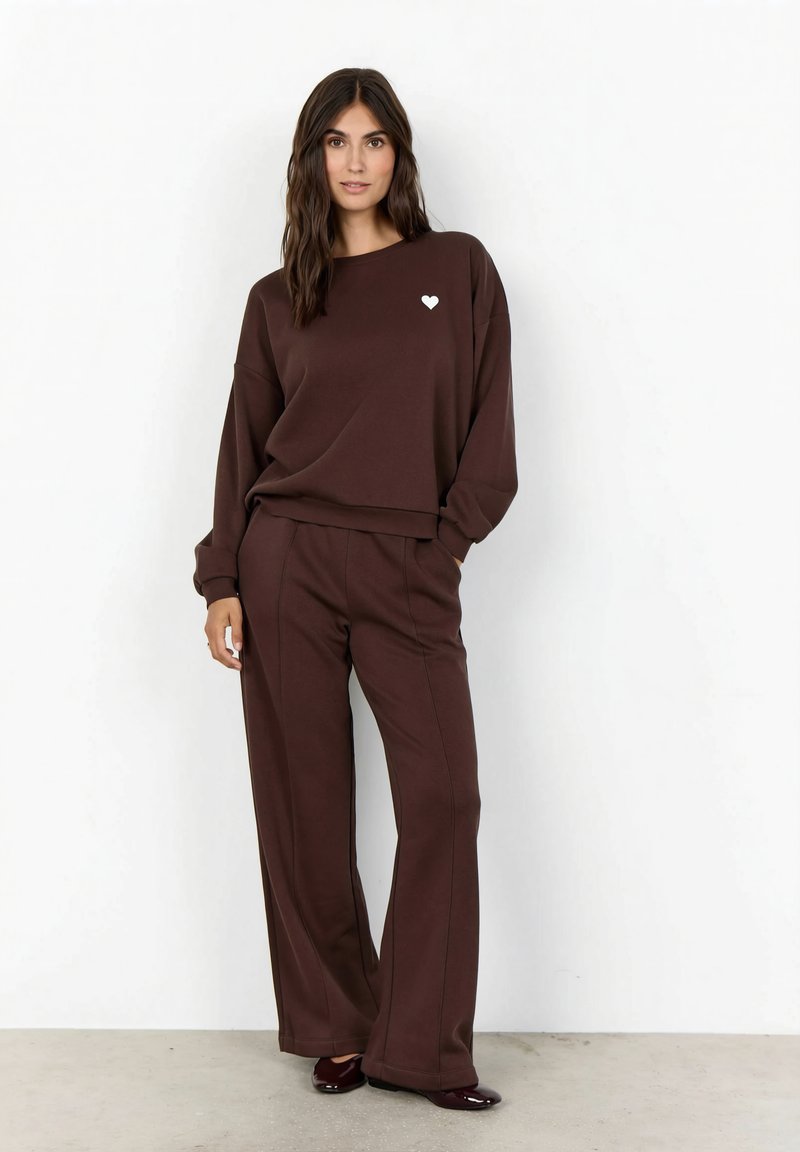 Femeie care poartă un hanorac asortat maro închis și pantaloni evazați cu o inimă albă mică pe piept, stând în fața unui perete alb.