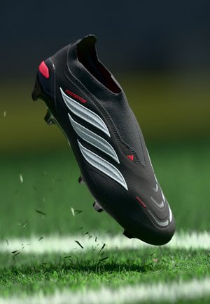 Melns futbola apavu ar sudraba svītrām, teksturētu virsmu un sarkanu akcentu. Raksturo ciešu piegulšanu un paceltas tapu zoles labākai saķerei.