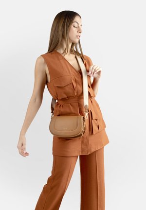 Donna che indossa un gilet senza maniche color ruggine e pantaloni abbinati, con una borsa a tracolla beige con una cinghia larga, che posa contro uno sfondo bianco.
