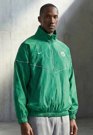 Mies, joka pukeutunut vihreään Nike-urheilutakkiin, jossa Boston Celticsin logo, seisoo yksinkertaisen betoniseinän edessä.