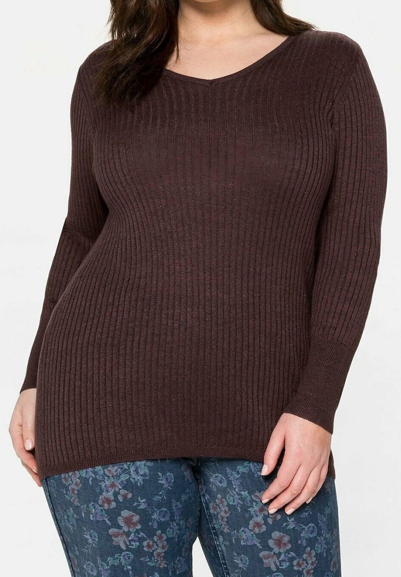 Femme portant un pull ajusté côtelé marron foncé à manches longues et un jean bleu à motif floral, debout la main près de la cuisse.
