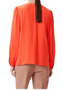 Blusa arancione brillante a maniche lunghe con una texture liscia, maniche arricciate e una leggera lucentezza. Abbinata a pantaloni a quadri marroni.