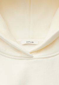 Hellcremefarbenes Kleidungsstück mit Kragen, das die Innenseite des Halsausschnitts zeigt, mit einem weißen OPUS Markenschild, das in der Mitte angenäht ist.