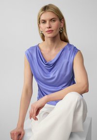 Bluzka z dekoltem typu cowl w jasnofioletowym kolorze, wykonana z gładkiego materiału. Krótkie rękawy, luźny krój; noszona z białymi spodniami. Elegancki design z minimalnymi akcentami.
