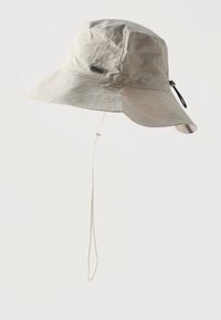 Ανοιχτό μπεζ καπέλο κουβά με φαρδύ γείσο, ρυθμιζόμενο λουρί κάτω από το σαγόνι και μικρή ετικέτα με το λογότυπο στην πλευρά.