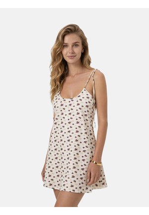 Donna con capelli biondi ondulati che indossa un vestito senza maniche con stampa floreale, spalline sottili e un braccialetto d'oro al polso destro.