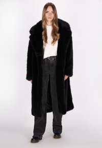 Lang sort faux fur frakke med en bred krave, båret over en hvid sweater og sorte højtaljede bukser med lige ben.