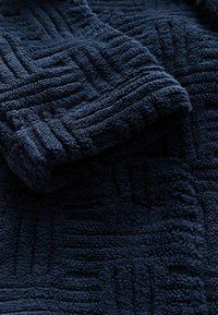 Granatowy, teksturowany koc z geometrycznym wzorem, wykonany z miękkiej, puszystej tkaniny oraz o pluszowej nawierzchni zapewniającej komfort i ciepło.