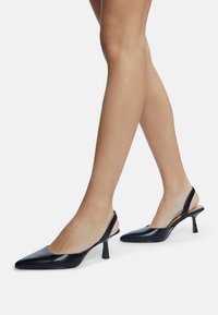 Jambes portant des chaussures à talons kitten à bout pointu et à bride arrière noires, avec un talon slim et anglé, sur un fond blanc.