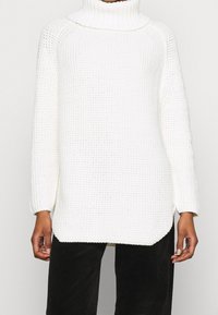 Biały dziany sweter z wysokim golfem, prążkowaną strukturą, luźnym krojem i długimi rękawami; boczne rozcięcia u dołu zwiększają komfort.