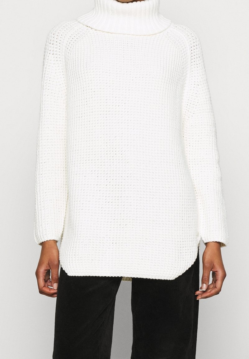 Biały dziany sweter z wysokim golfem, prążkowaną strukturą, luźnym krojem i długimi rękawami; boczne rozcięcia u dołu zwiększają komfort.
