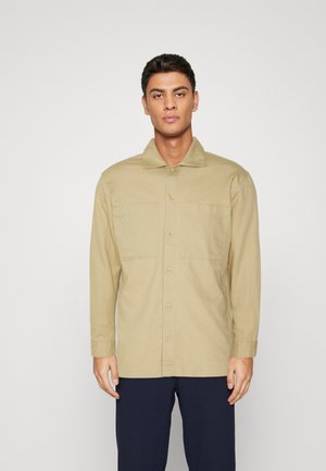 Beige Langarmhemd aus Baumwolle mit Kragen, Vorderknopfverschluss und zwei Brusttaschen, kombiniert mit dunklen Hosen.