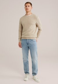 Mladic u bež džemperu, svijetloplavim trapericama i bijelim tenisicama, stoji naslonjen na jednostavnu svijetlu pozadinu.