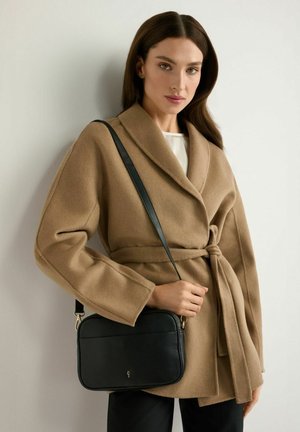 Kobieta w płaszczu w kolorze wielbłądzim, z paskiem, niosąca na ramieniu czarną skórzaną torbę na ramię, stoi na tle jednolitej ściany.