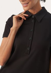 Persona sorridente che sistema il colletto di una polo nera a maniche corte con sei bottoni neri sul davanti.