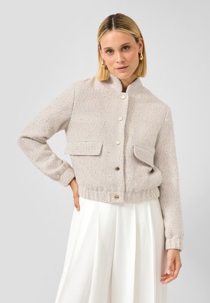 Femme portant une veste boutonnée beige clair texturée avec poches à rabat devant et un pantalon large blanc plissé, sur un fond uni.