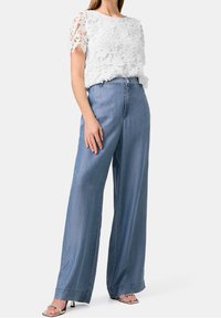 Kobieta w białej koronkowej bluzce z krótkim rękawem, w niebieskich dżinsowych spodniach z wysokim stanem i szerokimi nogawkami, oraz w srebrnych sandałach na obcasie, stojąca na białym tle.