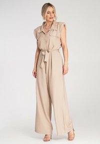 Beżowy kombinezon z krótkimi rękawami, kieszeniami z przodu, zapięciem na guziki oraz wiązanym paskiem w talii. Gładki materiał w luźnym kroju z szerokimi nogawkami.