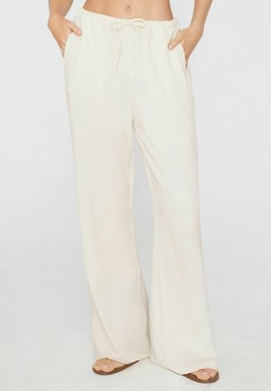 Femme portant un pantalon ample couleur crème, taille haute avec une taille à cordon et les mains dans les poches, associé à des tongs marron.