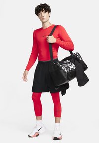 Rød langærmet sports T-shirt, sorte shorts, røde leggings, bærende en sort duffel taske med et hvidt og grått mønster. Hvite sneakers med lyserøde detaljer.