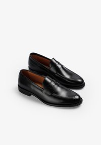 Mocasines de cuero negro con un acabado liso, que presentan una correa decorativa y detalles de costura. Suelas planas y forma de punta redonda.