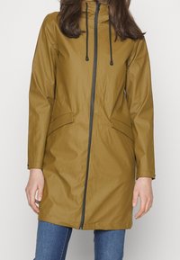 Giacca marrone resistente all'acqua con un alto colletto, chiusura con zip e due tasche laterali. Presenta lacci neri e una texture liscia.