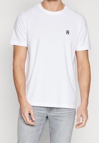 T-shirt bianco a maniche corte in cotone, con scollo rotondo e un piccolo logo scuro sul lato sinistro del petto. Texture morbida e liscia.