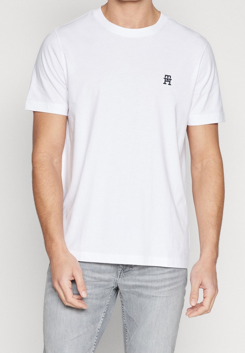 T-shirt bianco a maniche corte in cotone, con scollo rotondo e un piccolo logo scuro sul lato sinistro del petto. Texture morbida e liscia.
