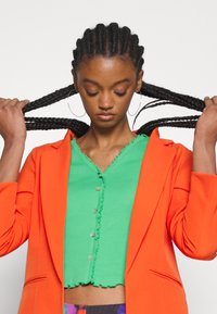 Fényes narancssárga zakó zöld, bordázott crop top felett, amelynek hullámos nyaka és gomb részlete van; a modell fonott hajat tart.