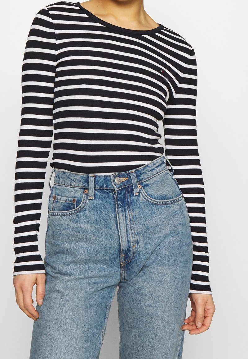 Camicia a righe bianche e nere con maniche lunghe abbinata a jeans azzurri a vita alta, caratterizzati da un taglio classico e un discreto logo sul petto.