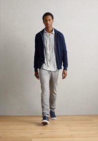 Un sweat à capuche zippé marine porté sur une chemise à rayures claires, associé à un pantalon gris clair et des baskets de sport noires, debout sur un sol en bois.