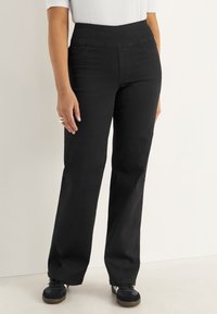 Nainen, jolla on yllään mustat korkeavyötäröiset suorat farkut ja mustat lenkkarit, valkoista taustaa vasten, kuvattu vyötäröstä ylöspäin ja polvista alaspäin.