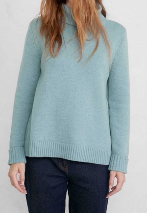 Person trägt einen hellblauen gestrickten Pullover mit umgeschlagenen Bündchen und dunkelblaue Jeans vor einem einfachen hellen Hintergrund.