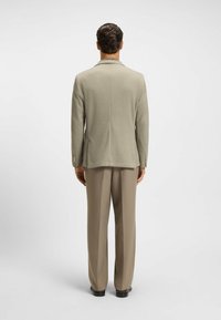 Hellgrauer Blazer aus strukturiertem Stoff, mit einem Schlitz hinten und paspelierten Revers, kombiniert mit beigen Hosen und schwarzen Schuhen.