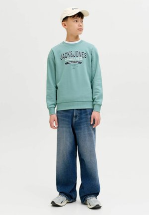 Poika, jolla on beige lippalakki, mintunvihreä Jack & Jones -collegepaita, leveät siniset farkut ja harmaat lenkkarit, seisomassa ja katsomassa sivulle.