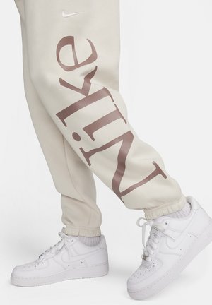 Pantalones de chándal beige con un gran texto en marrón "Nike" en la pierna, combinados con zapatillas blancas con perforaciones y un diseño clásico.