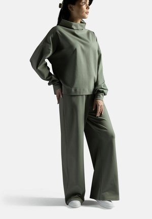 Nainen, jolla on yllään ylipitkä oliivinvihreä pooloneule ja leveälahkeiset housut sekä valkoiset tennarit, seisoo käsi lanteilla.