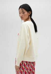 Felpa color crema con una vestibilità rilassata, polsini a coste e scollo rotondo. Indossata con pantaloni a fantasia rossi, rosa e beige.