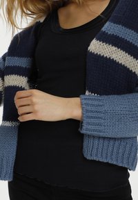 Kvinna som bär en blårandig stickad cardigan över en svart ribbad ärmlös topp, hållande cardiganen öppen med ena handen.