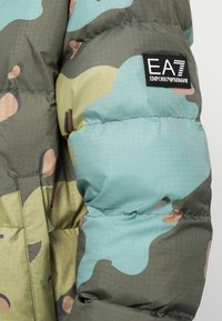 Kamuflaažiga puffernahk, millel on rohelise, sinise ja beeži mustriga segu. Omab tikandit ja musta logo plaati. Pehme kangas.