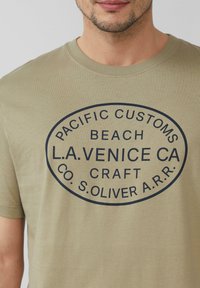 Mężczyzna w jasnooliwkowej koszulce z granatowym owalnym logo, na którym widnieje napis "Pacific Customs Beach L.A. Venice CA Craft Co. S. Oliver A.R.R."